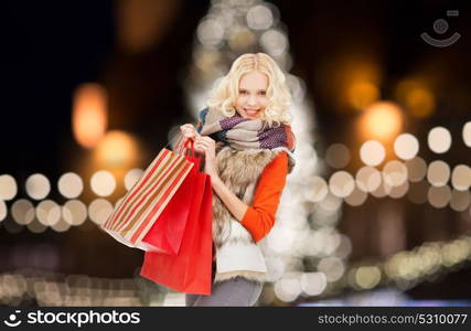 sale, holidays and people concept - happy teenage girl or young woman in winter clothes with shopping bags over christmas tree lights background. young woman in winter clothes with shopping bags