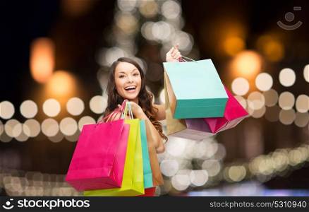 sale, holidays and people concept - smiling woman in red dress with colorful shopping bags over christmas tree lights background. woman in red dress with colorful shopping bags