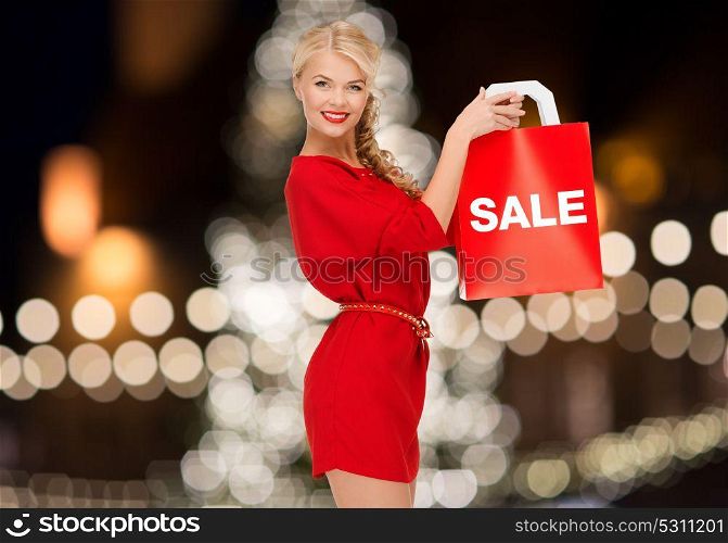 sale, holidays and people concept - smiling woman in red dress with shopping bag over christmas tree lights background. woman in red dress with word sale on shopping bag