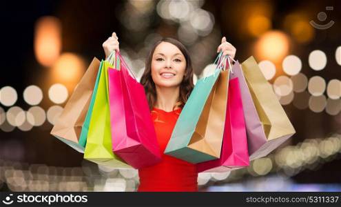 sale, holidays and people concept - smiling woman in red dress with colorful shopping bags over christmas tree lights background. woman in red dress with colorful shopping bags