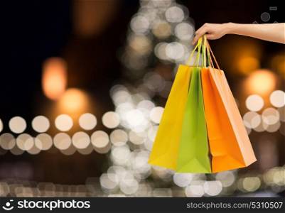 sale, holidays, consumerism and people concept - close up of hand holding shopping bags over christmas tree lights background. close up of hand with shopping bags at christmas