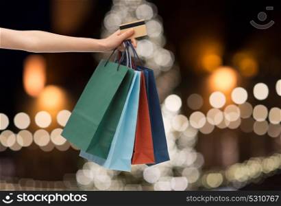 sale, holidays, consumerism and people concept - close up of hand holding shopping bags and credit card over christmas tree lights background. shopping bags and credit card in hand at christmas