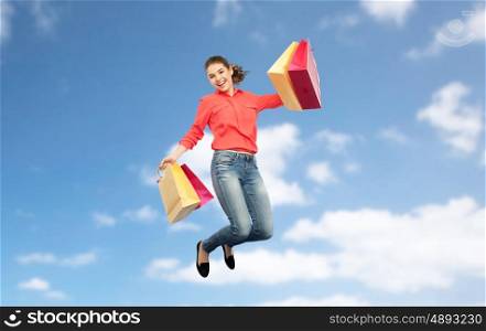 sale, motion and people concept - smiling young woman with shopping bags jumping in air over blue sky and clouds background
