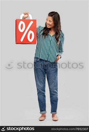 sale, outlet and consumerism concept - happy asian young woman with percentage sign on red shopping bag over grey background. asian woman with percentage sign on shopping bags