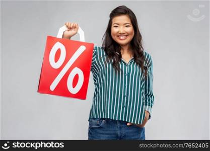 sale, outlet and consumerism concept - happy asian young woman with percentage sign on red shopping bag over grey background. asian woman with percentage sign on shopping bags