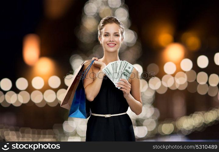 sale, people and holidays concept - smiling woman in dress with shopping bags and money over christmas tree lights background. woman with shopping bags and money at christmas