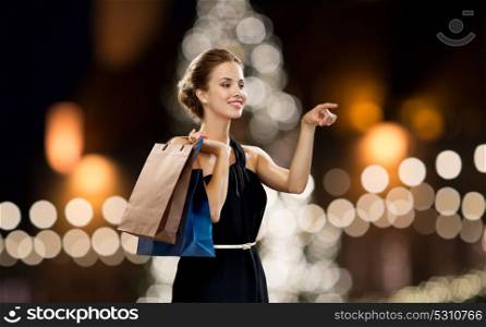 sale, people and luxury concept - happy beautiful young woman in black dress with shopping bags over christmas tree lights background. woman in black with shopping bags at christmas