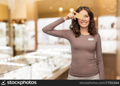 sale, shopping and business concept - happy female shop assistant covering her eye with golden credit card over jewelry store background. happy female shop assistant with credit card