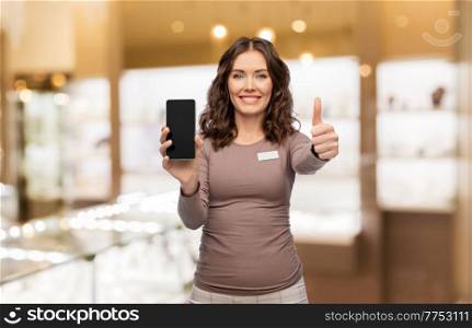 sale, shopping and business concept - happy female shop assistant with name tag showing smartphone over jewelry store background. happy female shop assistant showing smartphone