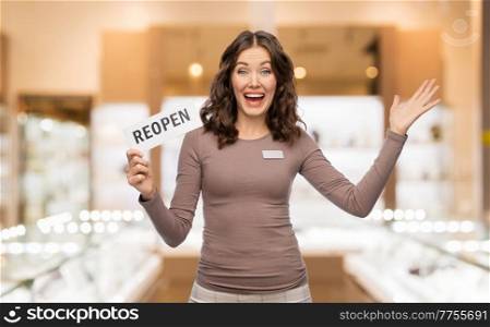 sale, shopping and business concept - happy female shop assistant with reopen sign and name tag over jewelry store background. happy jewelry shop assistant with reopen sign