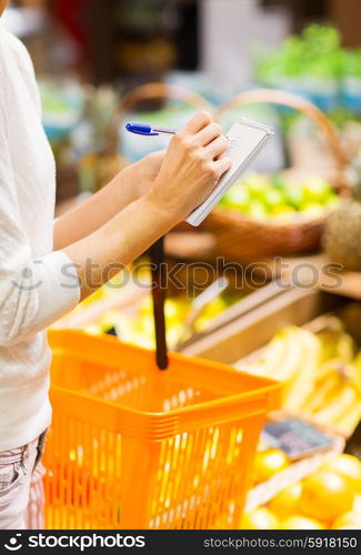 sale, shopping, consumerism and people concept - close up of young woman with basket taking notes to notebook in market