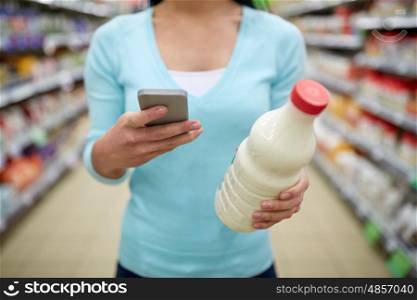 sale, shopping, consumerism and people concept - young woman with smartphone holding milk bottle at grocery store or supermarket