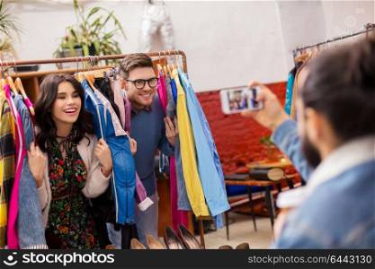 sale, shopping, fashion and people concept - friend photographing happy couple having fun by smartphone at vintage clothing store hanger. happy couple having fun at vintage clothing store