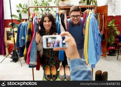 sale, shopping, fashion and people concept - friend photographing happy couple having fun by smartphone at vintage clothing store hanger. friend photographing couple at clothing store