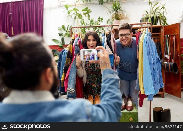 sale, shopping, fashion and people concept - friend photographing happy couple having fun by smartphone at vintage clothing store hanger. friend photographing couple at clothing store. friend photographing couple at clothing store