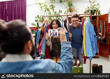 sale, shopping, fashion and people concept - friend photographing happy couple having fun by smartphone at vintage clothing store hanger. friend photographing couple at clothing store. friend photographing couple at clothing store