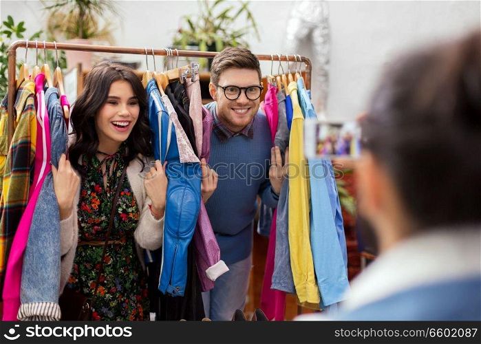 sale, shopping, fashion and people concept - friend photographing happy couple having fun by smartphone at vintage clothing store hanger. happy couple having fun at vintage clothing store