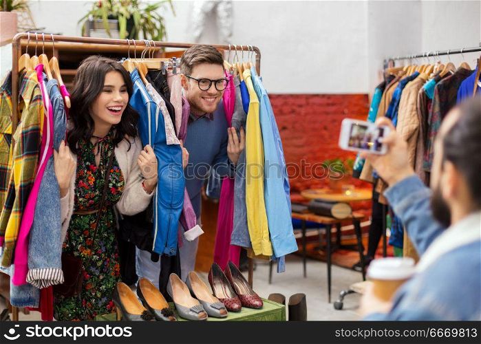 sale, shopping, fashion and people concept - friend photographing happy couple having fun by smartphone at vintage clothing store hanger. happy couple having fun at vintage clothing store. happy couple having fun at vintage clothing store