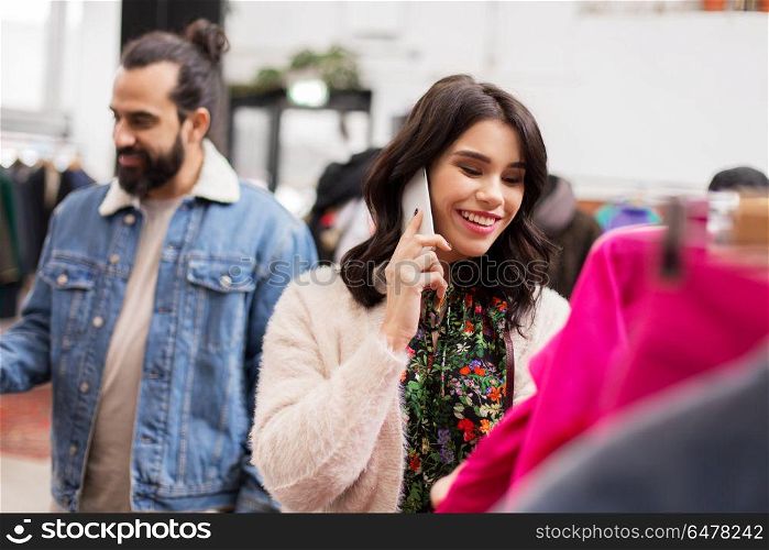 sale, shopping, fashion and people concept - happy young woman calling on smartphone and choosing clothes at vintage clothing store. woman calling on smartphone at clothing store. woman calling on smartphone at clothing store