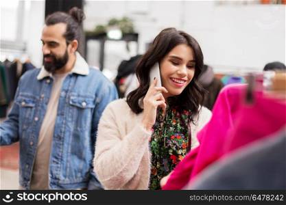 sale, shopping, fashion and people concept - happy young woman calling on smartphone and choosing clothes at vintage clothing store. woman calling on smartphone at clothing store. woman calling on smartphone at clothing store