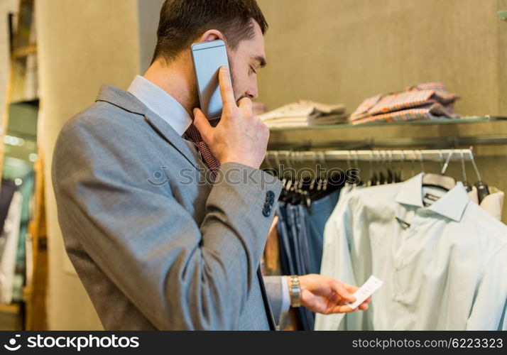 sale, shopping, fashion, communication and people concept - close up of young man calling on smartphone and looking to shirt price tag at clothing store