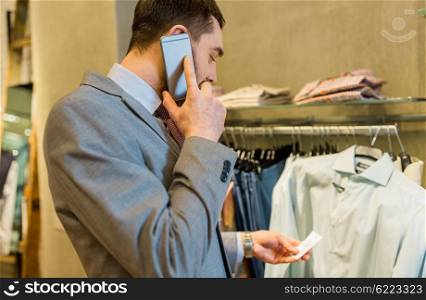 sale, shopping, fashion, communication and people concept - close up of young man calling on smartphone and looking to shirt price tag at clothing store