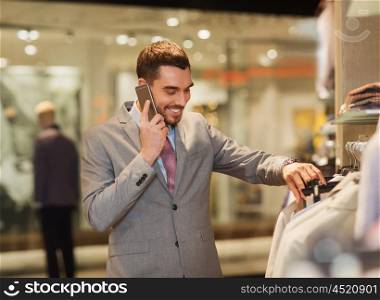 sale, shopping, fashion, communication and people concept - happy young man calling on smartphone and choosing clothes at clothing store