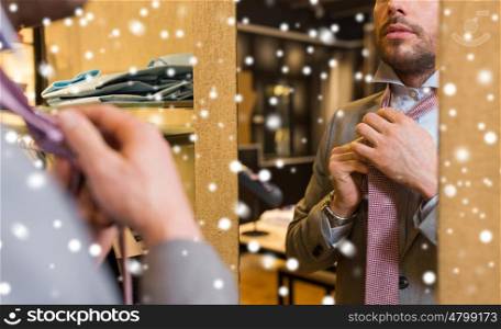sale, shopping, fashion, style and people concept - close up of young man in suit choosing and tying tie and looking to mirror in mall or clothing store over snow