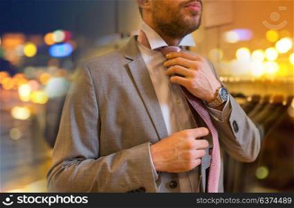 sale, shopping, fashion, style and people concept - close up of young man in suit choosing and tying tie in mall or clothing store. close up of man tying tie at clothing store