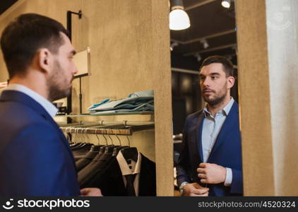 sale, shopping, fashion, style and people concept - elegant young man choosing and trying jacket on and looking to mirror in mall or clothing store