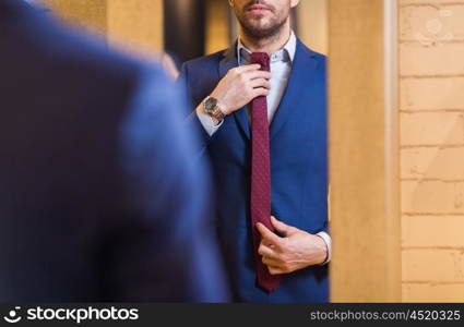 sale, shopping, fashion, style and people concept - elegant young man choosing and trying tie on and looking to mirror in mall or clothing store
