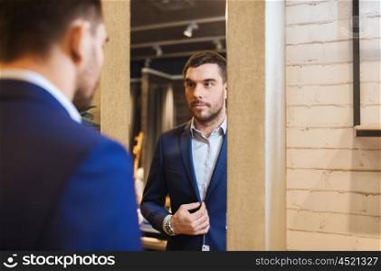 sale, shopping, fashion, style and people concept - elegant young man choosing and trying jacket on and looking to mirror in mall or clothing store