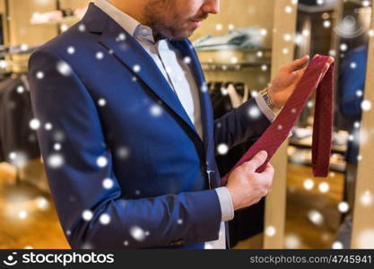 sale, shopping, fashion, style and people concept - elegant young man choosing and trying tie on at clothing store over snow