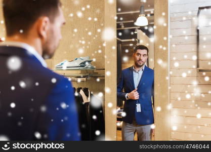 sale, shopping, fashion, style and people concept - elegant young man choosing and trying jacket on and looking to mirror in mall or clothing store over snow