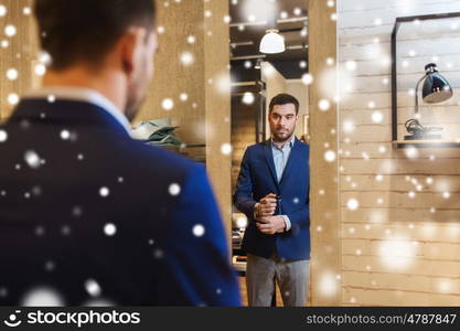 sale, shopping, fashion, style and people concept - elegant young man choosing and trying jacket on and looking to mirror in mall or clothing store over snow