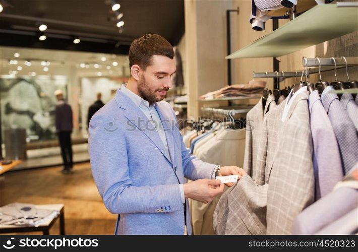 sale, shopping, fashion, style and people concept - elegant young man in jacket choosing clothes and looking at price tag in mall or clothing store