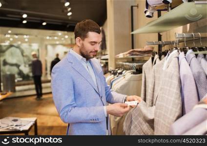 sale, shopping, fashion, style and people concept - elegant young man in jacket choosing clothes and looking at price tag in mall or clothing store