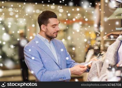 sale, shopping, fashion, style and people concept - elegant young man in jacket choosing clothes and looking at price tag in mall or clothing store over snow