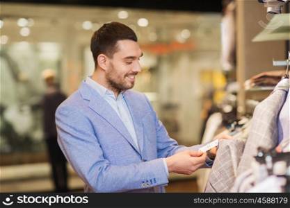 sale, shopping, fashion, style and people concept - happy elegant young man in jacket choosing clothes and looking at price tag in mall or clothing store