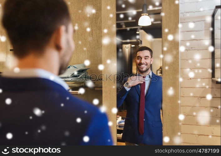 sale, shopping, fashion, style and people concept - happy young man choosing and trying tie on and looking to mirror in mall or clothing store over snow