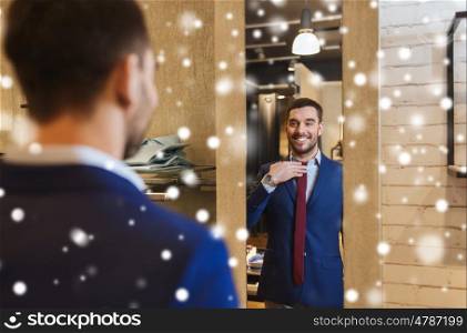 sale, shopping, fashion, style and people concept - happy young man choosing and trying tie on and looking to mirror in mall or clothing store over snow