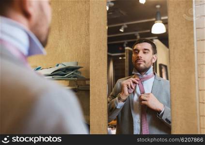 sale, shopping, fashion, style and people concept - happy young man choosing and tying tie on and looking to mirror in mall or clothing store