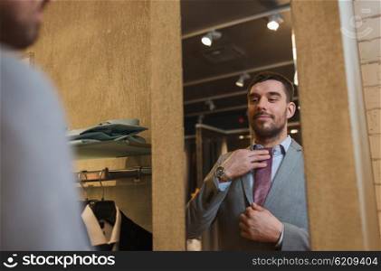 sale, shopping, fashion, style and people concept - happy young man choosing and trying tie on and looking to mirror in mall or clothing store