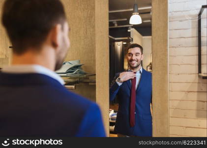 sale, shopping, fashion, style and people concept - happy young man choosing and trying tie on and looking to mirror in mall or clothing store