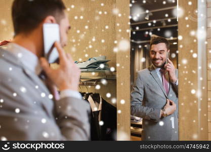 sale, shopping, fashion, style and people concept - happy young man or businessman trying suit and tie on and calling on smartphone at clothing store mirror over snow