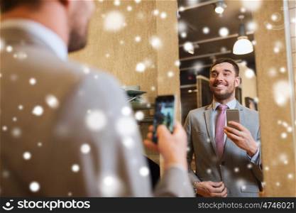 sale, shopping, fashion, style and people concept - happy young man or businessman in suit with smartphone taking mirror selfie at clothing store over snow