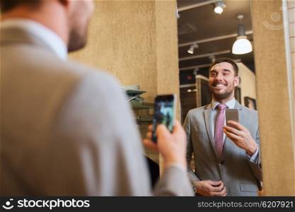 sale, shopping, fashion, style and people concept - happy young man or businessman in suit with smartphone taking mirror selfie at clothing store