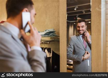 sale, shopping, fashion, style and people concept - happy young man or businessman trying suit and tie on and calling on smartphone at clothing store mirror