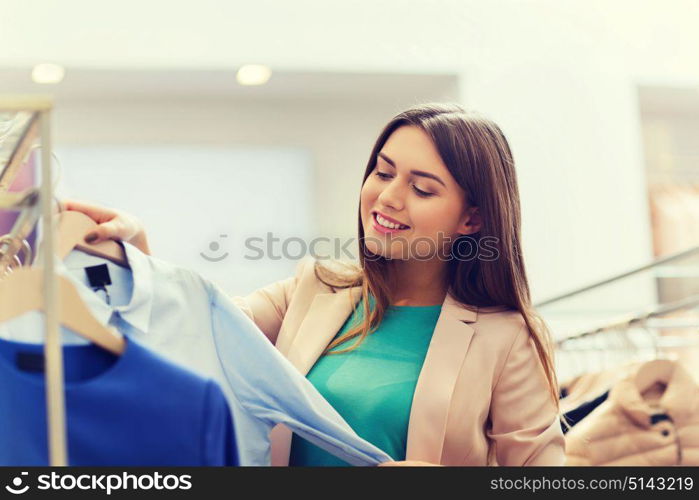 sale, shopping, fashion, style and people concept - happy young woman choosing clothes in mall or clothing store. happy young woman choosing clothes in mall