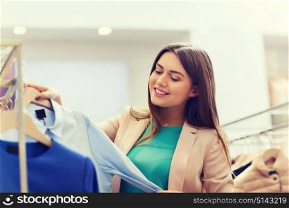 sale, shopping, fashion, style and people concept - happy young woman choosing clothes in mall or clothing store. happy young woman choosing clothes in mall